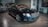 A sleek, luxurious black car with shiny chrome details is parked indoors. The vehicle features a prominent front grille and distinctive headlights. There are other cars present in the background, suggesting a showroom or garage setting. The tiled floor and wall art add to the upscale ambiance.