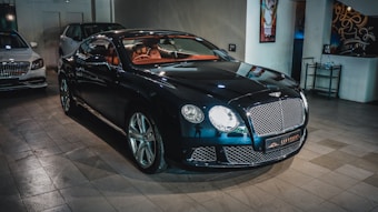 A sleek, luxurious black car with shiny chrome details is parked indoors. The vehicle features a prominent front grille and distinctive headlights. There are other cars present in the background, suggesting a showroom or garage setting. The tiled floor and wall art add to the upscale ambiance.