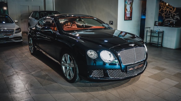 A sleek, luxurious black car with shiny chrome details is parked indoors. The vehicle features a prominent front grille and distinctive headlights. There are other cars present in the background, suggesting a showroom or garage setting. The tiled floor and wall art add to the upscale ambiance.