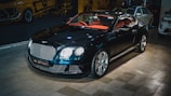 A luxury sports car with a glossy black finish is parked indoors. The vehicle features a stylish red interior and sleek design elements. The background includes automotive-themed wall art and two other cars are partially visible. The car is prominently displayed with its chrome accents and distinctive grille.