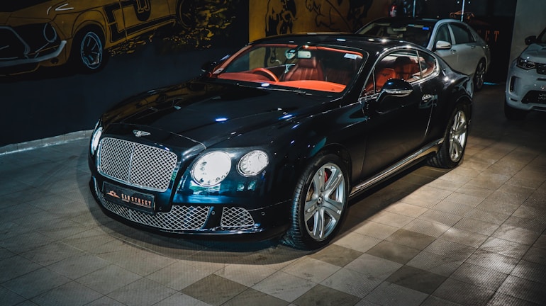 A luxury sports car with a glossy black finish is parked indoors. The vehicle features a stylish red interior and sleek design elements. The background includes automotive-themed wall art and two other cars are partially visible. The car is prominently displayed with its chrome accents and distinctive grille.