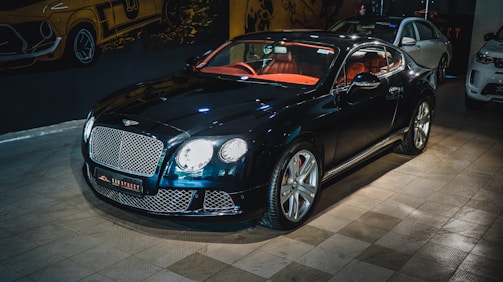 A luxury sports car with a glossy black finish is parked indoors. The vehicle features a stylish red interior and sleek design elements. The background includes automotive-themed wall art and two other cars are partially visible. The car is prominently displayed with its chrome accents and distinctive grille.