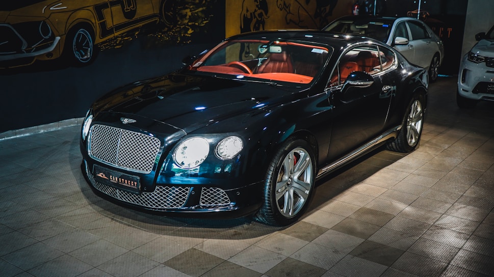 A luxury sports car with a glossy black finish is parked indoors. The vehicle features a stylish red interior and sleek design elements. The background includes automotive-themed wall art and two other cars are partially visible. The car is prominently displayed with its chrome accents and distinctive grille.