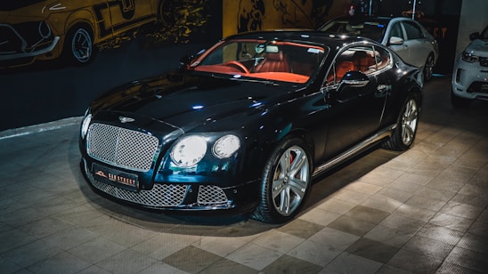 A luxury sports car with a glossy black finish is parked indoors. The vehicle features a stylish red interior and sleek design elements. The background includes automotive-themed wall art and two other cars are partially visible. The car is prominently displayed with its chrome accents and distinctive grille.