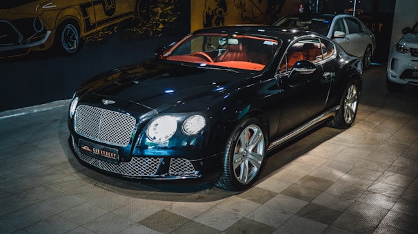 A luxury sports car with a glossy black finish is parked indoors. The vehicle features a stylish red interior and sleek design elements. The background includes automotive-themed wall art and two other cars are partially visible. The car is prominently displayed with its chrome accents and distinctive grille.