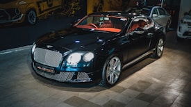 A luxury sports car with a glossy black finish is parked indoors. The vehicle features a stylish red interior and sleek design elements. The background includes automotive-themed wall art and two other cars are partially visible. The car is prominently displayed with its chrome accents and distinctive grille.