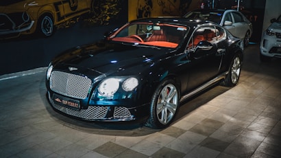 A luxury sports car with a glossy black finish is parked indoors. The vehicle features a stylish red interior and sleek design elements. The background includes automotive-themed wall art and two other cars are partially visible. The car is prominently displayed with its chrome accents and distinctive grille.