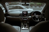 The interior of a modern car featuring a sleek dashboard design with an infotainment system, climate controls, and a leather-wrapped steering wheel. The front seats are visible, and the view through the windshield shows another car passing by on the road outside.