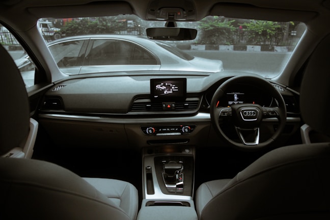The interior of a modern car featuring a sleek dashboard design with an infotainment system, climate controls, and a leather-wrapped steering wheel. The front seats are visible, and the view through the windshield shows another car passing by on the road outside.