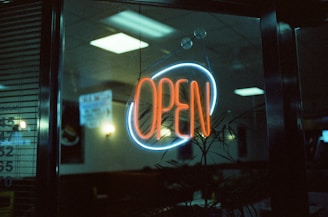 A neon sign displaying the word 'open' in vibrant orange and blue hues, hanging inside a glass window of what appears to be a diner or restaurant. The dimly lit interior includes some visible furniture, and the reflection of ceiling lights can be seen on the window.