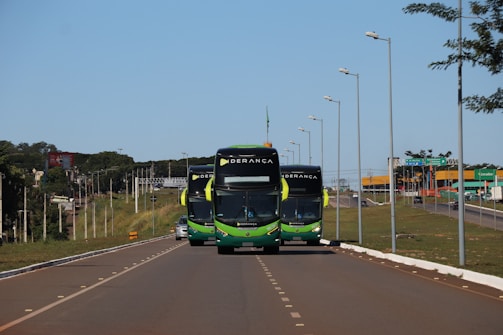 Three buses with a green and black color scheme travel along a well-maintained highway. Street lamps line both sides of the road. In the background, there is a green landscape with sparse trees and a blue sky.