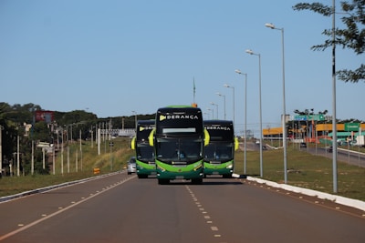 Three buses with a green and black color scheme travel along a well-maintained highway. Street lamps line both sides of the road. In the background, there is a green landscape with sparse trees and a blue sky.