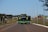 Three buses with a green and black color scheme travel along a well-maintained highway. Street lamps line both sides of the road. In the background, there is a green landscape with sparse trees and a blue sky.
