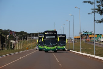 Three buses with a green and black color scheme travel along a well-maintained highway. Street lamps line both sides of the road. In the background, there is a green landscape with sparse trees and a blue sky.
