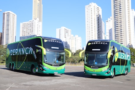 Two large, modern buses with a sleek green and black design featuring the word 'LIDERANÇA' prominently on the side are parked in an urban setting. The background consists of tall, white high-rise buildings and some greenery.