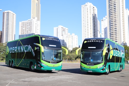 Two large, modern buses with a sleek green and black design featuring the word 'LIDERANÇA' prominently on the side are parked in an urban setting. The background consists of tall, white high-rise buildings and some greenery.