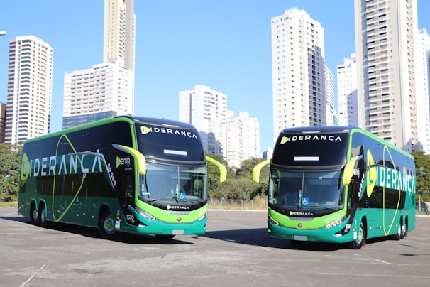 Two large, modern buses with a sleek green and black design featuring the word 'LIDERANÇA' prominently on the side are parked in an urban setting. The background consists of tall, white high-rise buildings and some greenery.