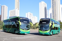 Two large, modern buses with a sleek green and black design featuring the word 'LIDERAN&Ccedil;A' prominently on the side are parked in an urban setting. The background consists of tall, white high-rise buildings and some greenery.