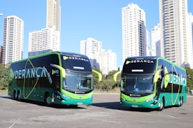 Two large, modern buses with a sleek green and black design featuring the word 'LIDERANÇA' prominently on the side are parked in an urban setting. The background consists of tall, white high-rise buildings and some greenery.