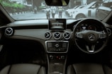 The interior of a luxury car featuring a sleek dashboard with a center console screen. Prominent circular air vents and a multifunction steering wheel are visible. Through the windshield, a street view with a few pedestrians and parked cars is noticeable.