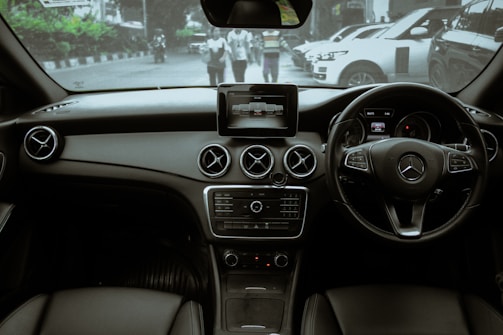 The interior of a luxury car featuring a sleek dashboard with a center console screen. Prominent circular air vents and a multifunction steering wheel are visible. Through the windshield, a street view with a few pedestrians and parked cars is noticeable.