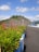 A paved road with blue and white barriers runs alongside neatly trimmed bushes. In the background, a large, multi-level embankment covered in green foliage rises up, with 'Bendungan Wonorejo' written on it. The sky is bright and dotted with fluffy clouds.