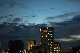 High-rise buildings are silhouetted against a dark blue evening sky, with scattered clouds creating a dramatic effect. The largest building displays lit windows and a prominent letter 'R' near the top.