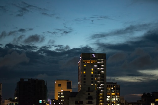 High-rise buildings are silhouetted against a dark blue evening sky, with scattered clouds creating a dramatic effect. The largest building displays lit windows and a prominent letter 'R' near the top.