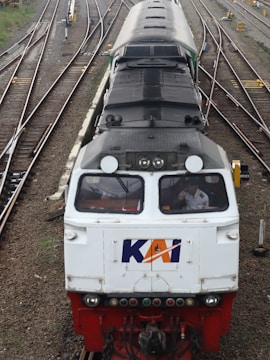 A train locomotive on railway tracks, viewed from above, with multiple tracks converging and diverging in the background. The front of the train is white with a logo, and a person is visible inside the driver's cabin.