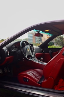 The interior of a luxury sports car with red leather seats and a sleek dashboard is visible. A steering wheel with a recognizable logo, a manual gear shift lever, and various controls and dials are present. The windows reveal a blurred view of urban buildings and greenery outside.