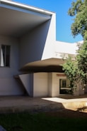 A modern architectural structure features angular overhangs and sharp lines. The building is predominantly white with a minimalist design. Partially visible is a sign indicating 'ESOLA SUPER' indicative of a school or educational institution. There are large glass windows and lush greenery surrounding the structure. The sky is clear and blue, enhancing the clean aesthetic of the building.