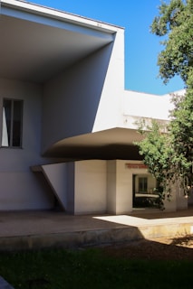 A modern architectural structure features angular overhangs and sharp lines. The building is predominantly white with a minimalist design. Partially visible is a sign indicating 'ESOLA SUPER' indicative of a school or educational institution. There are large glass windows and lush greenery surrounding the structure. The sky is clear and blue, enhancing the clean aesthetic of the building.