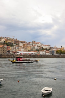 A picturesque riverside view of a city with colorful historic buildings stacked on a hill. A traditional boat is sailing on the river in the foreground, carrying passengers along the scenic route. The sky is partly cloudy, adding a serene backdrop to the lively scene on the water.