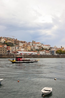 A picturesque riverside view of a city with colorful historic buildings stacked on a hill. A traditional boat is sailing on the river in the foreground, carrying passengers along the scenic route. The sky is partly cloudy, adding a serene backdrop to the lively scene on the water.