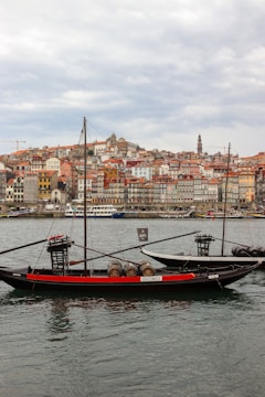 Two traditional Portuguese rabelo boats are anchored on a river, each carrying wooden barrels. In the background, a historic cityscape with colorful buildings and a prominent church tower rises atop a hill, under a cloudy sky.
