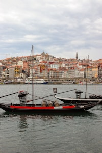 Two traditional Portuguese rabelo boats are anchored on a river, each carrying wooden barrels. In the background, a historic cityscape with colorful buildings and a prominent church tower rises atop a hill, under a cloudy sky.