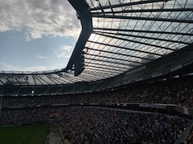 A large sports stadium filled with a crowd of spectators. The stadium has a modern, curved roof structure, with beams and glass panels, allowing natural light to filter through. People are seated in rows on expansive seating areas. The playing field is partially visible with goalposts, and the sky is partly cloudy.