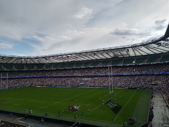 A large stadium filled with spectators, with a rugby game taking place on the field. The players are grouped near the center, possibly in a scrum. The stands are full, indicating a high attendance, and the sky is overcast with some patches of blue.