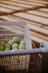 A plastic basket filled with tennis balls in varying states of wear sits on a surface with sunlight casting striped shadows across the scene.