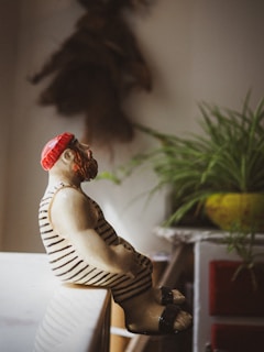 A ceramic figurine of a person wearing a red cap and a striped outfit sits on the edge of a surface. The surroundings include a plant with long, green leaves and a blurred background that suggests a cozy indoor setting.