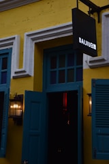 A yellow building facade with white-trimmed windows and blue shutters. A black sign with the word 'BAIJIU' in white is prominently displayed. Two wall-mounted lanterns are visible, casting a warm glow.