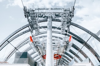 A close-up view of a modern cable car station, featuring intricate metal structures and supporting cables. The architecture includes large, curved glass panels and multiple visible layers of metalwork. The sky is partly cloudy, and there are pops of red color indicating caution or safety markers.