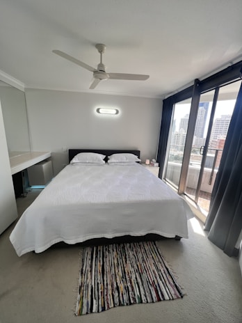 A modern bedroom with a neatly made double bed covered with a white bedspread. Above the bed, a modern light fixture is mounted on the wall. A small multicolored rug is placed on the floor at the foot of the bed. A ceiling fan is installed in the center of the room's ceiling. Large sliding glass doors with dark curtains open to a balcony with a view of city buildings.