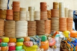 Stacks of spools of yarn are arranged in an orderly manner. The yarns vary in color, mainly in earthy tones like browns, oranges, and beige. Below the yarn spools, brightly colored synthetic ropes and strings are neatly coiled, featuring hues of red, green, yellow, and blue. Transparent tapes are stacked among the yarns.
