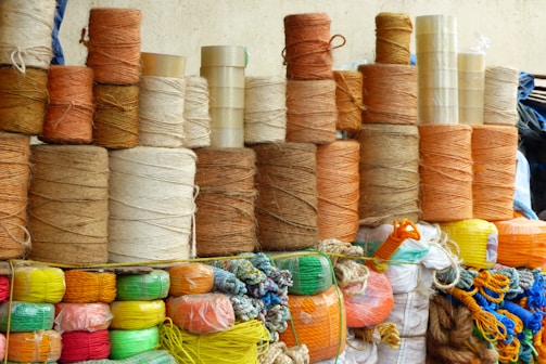 Stacks of spools of yarn are arranged in an orderly manner. The yarns vary in color, mainly in earthy tones like browns, oranges, and beige. Below the yarn spools, brightly colored synthetic ropes and strings are neatly coiled, featuring hues of red, green, yellow, and blue. Transparent tapes are stacked among the yarns.