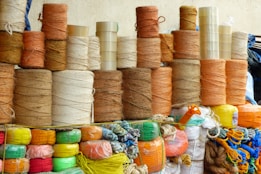 Stacks of spools of yarn are arranged in an orderly manner. The yarns vary in color, mainly in earthy tones like browns, oranges, and beige. Below the yarn spools, brightly colored synthetic ropes and strings are neatly coiled, featuring hues of red, green, yellow, and blue. Transparent tapes are stacked among the yarns.