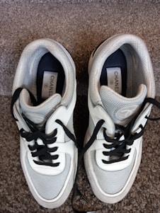A pair of stylish sneakers with white, black, and gray detailing is laid out on a carpeted floor. The shoes have black laces and feature a label with branding on the inside.