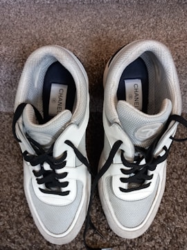 A pair of stylish sneakers with white, black, and gray detailing is laid out on a carpeted floor. The shoes have black laces and feature a label with branding on the inside.