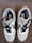 A pair of stylish sneakers with white, black, and gray detailing is laid out on a carpeted floor. The shoes have black laces and feature a label with branding on the inside.