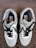 A pair of stylish sneakers with white, black, and gray detailing is laid out on a carpeted floor. The shoes have black laces and feature a label with branding on the inside.